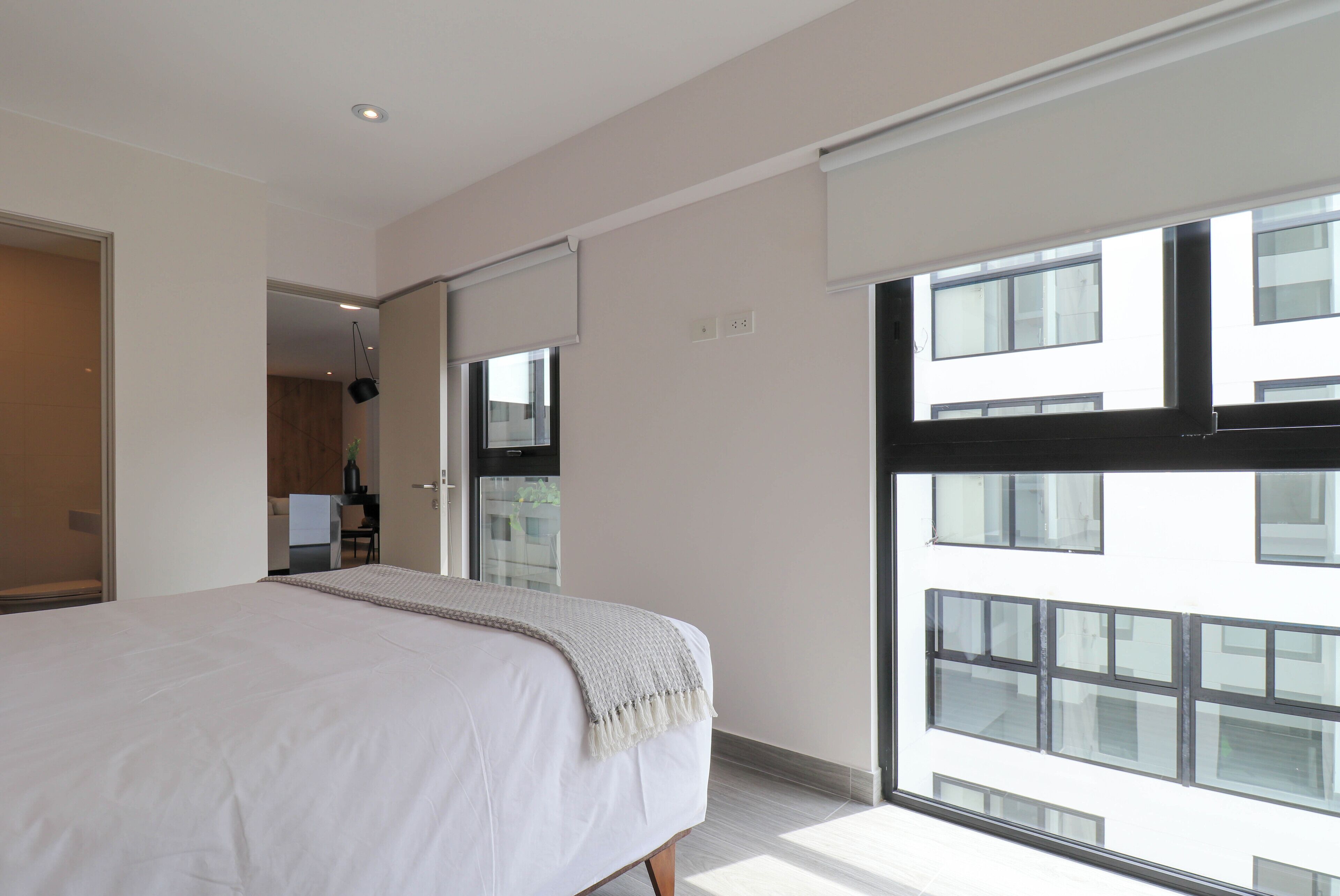Une chambre moderne avec un grand lit, une porte coulissante donnant sur une salle de bain et une vue sur un immeuble à travers de grandes fenêtres.