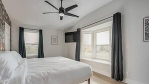 Quarto com cama, ventilador de teto, janela com cortinas e televisão fixada na parede.