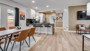 Uma cozinha moderna com banquetas, uma sala de jantar com mesa e cadeiras, e uma TV na parede.