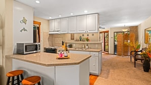 A spacious kitchen with white cabinets, a large island with stools, a microwave, and a wine bottle. - Waikomo Stream Villas 100 | Tropical Poipu Retreat Near Beach (Koloa)