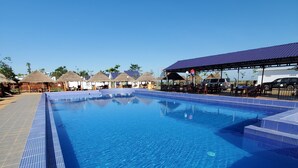 A clear blue swimming pool with tiled edges, surrounded by thatched-roof structures and a covered seating area. - I AM Koh Ker (Kuleaen)