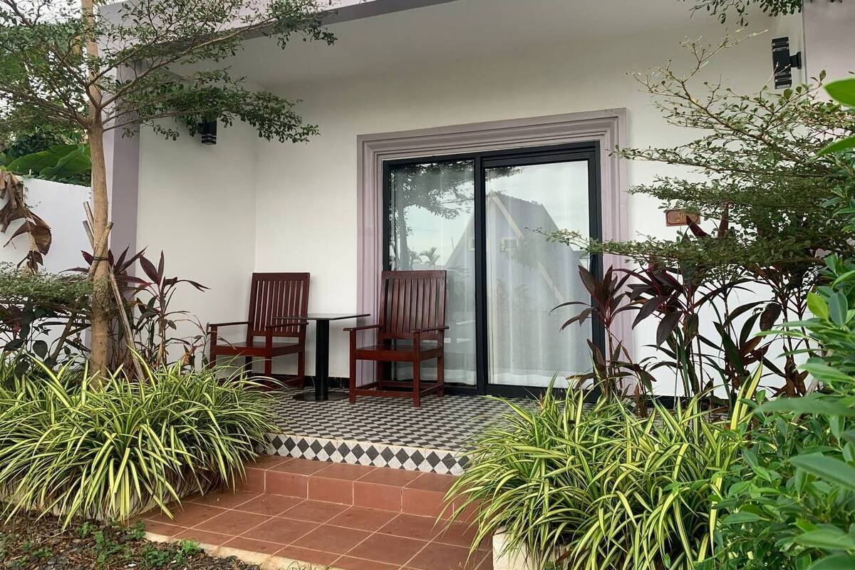 A covered outdoor seating area with two wooden chairs and a table, surrounded by potted plants and a tiled floor.