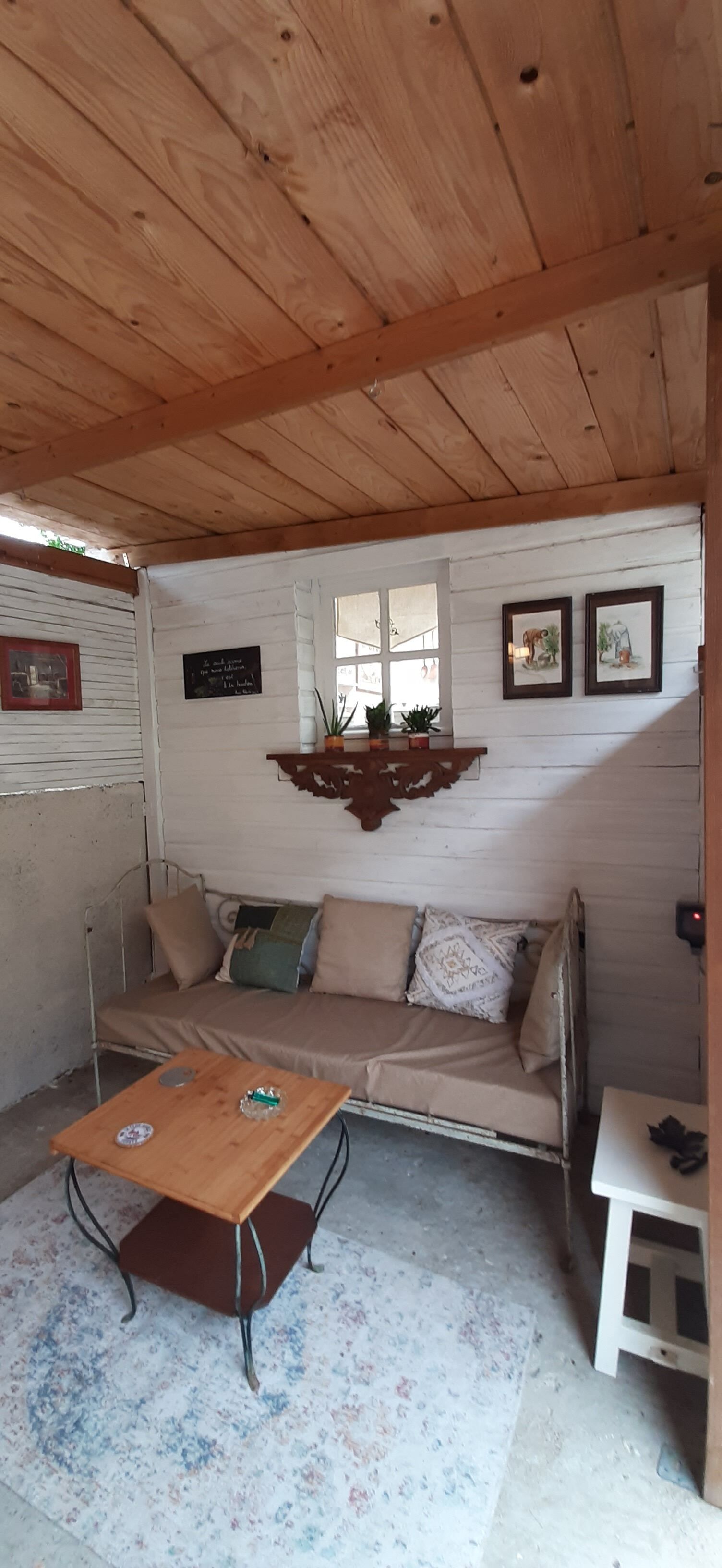 Een gezellige kamer met een houten plafond, een bank met kussens, een klein tafeltje en een raam met planten.