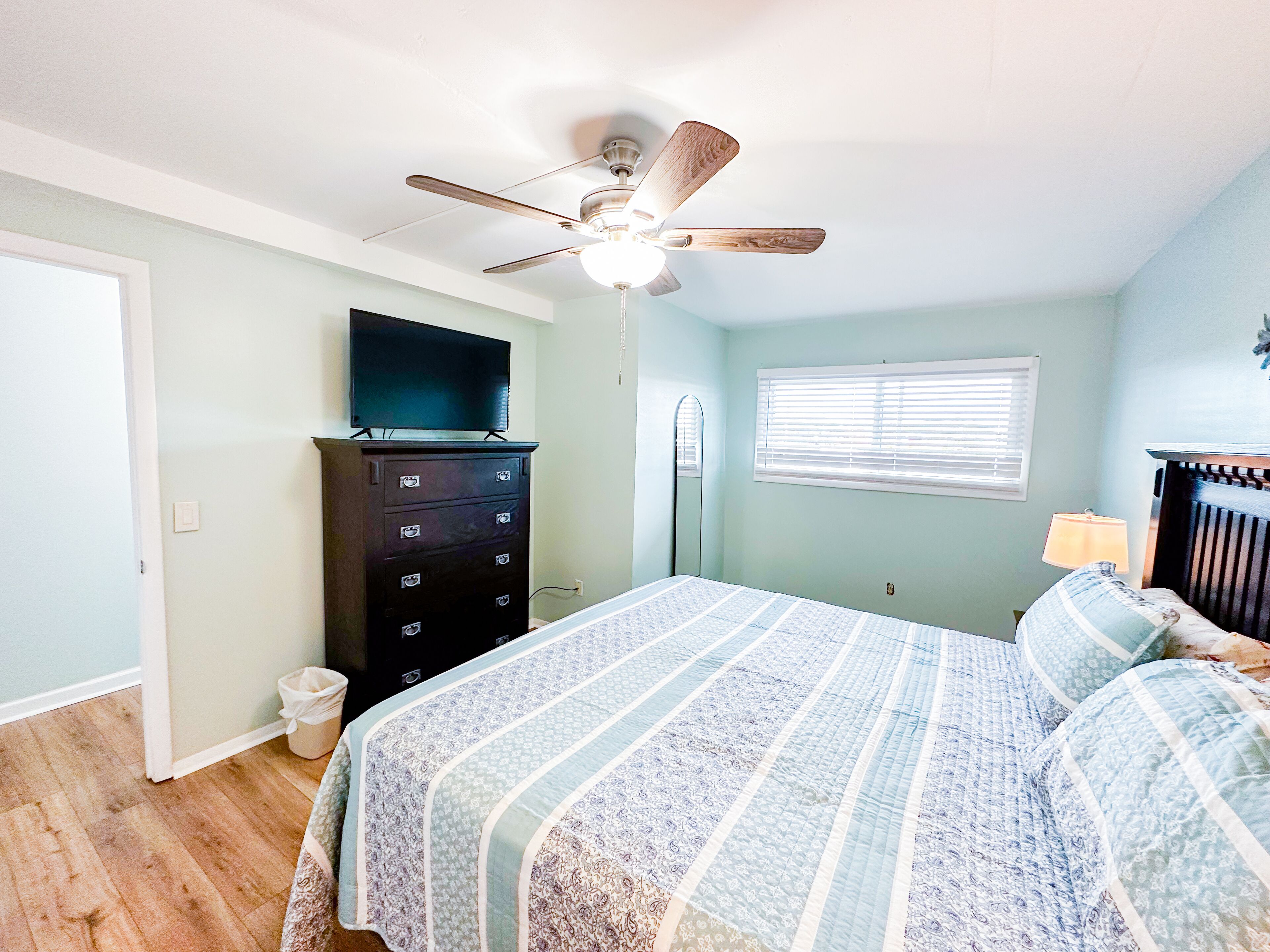 A bedroom with a bed, a dresser, a ceiling fan, a TV, a window with blinds, and a nightstand with a lamp.