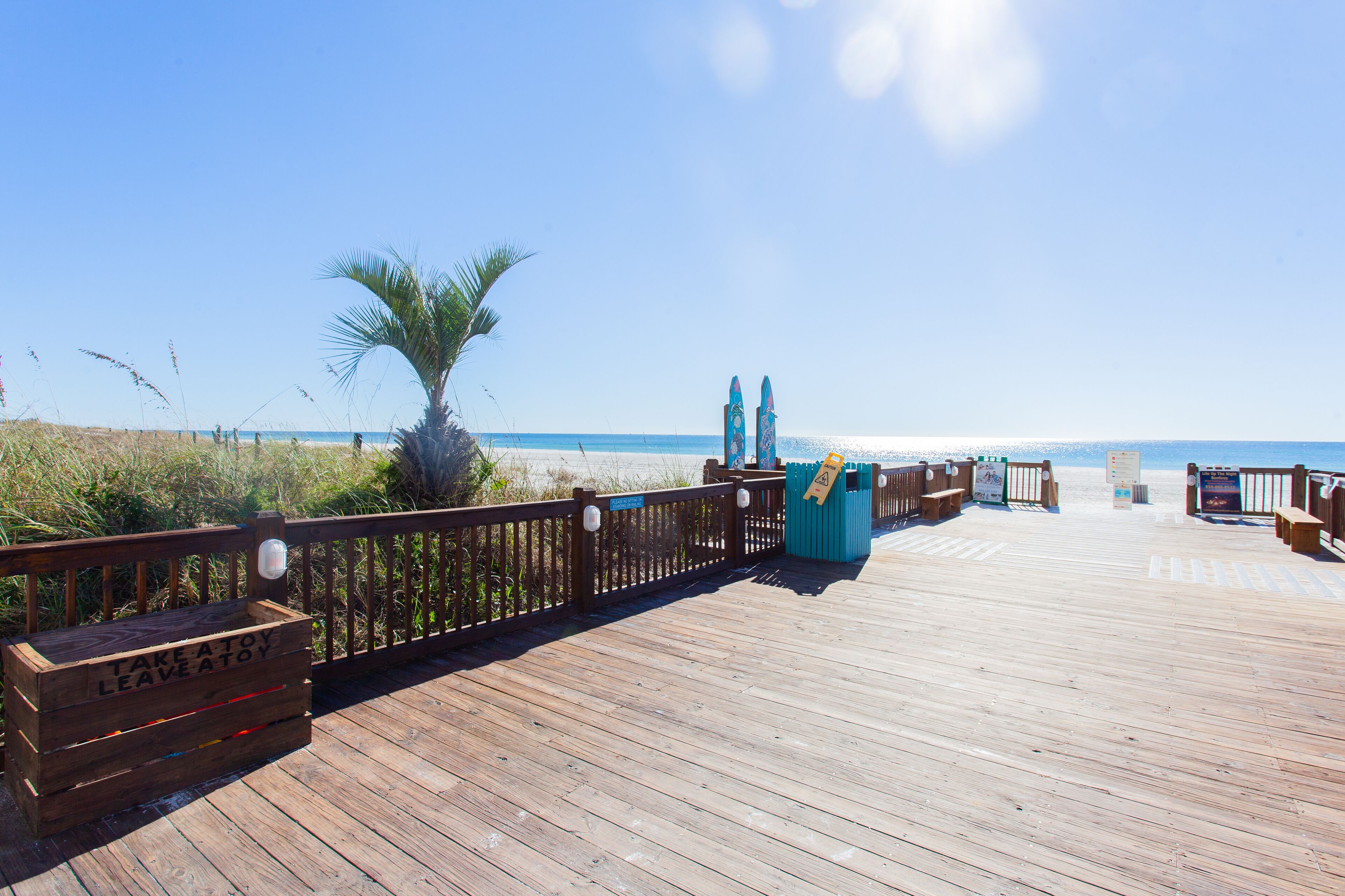 A wooden boardwalk leading to a beach with a palm tree and a sign that says "Take a toy, leave a toy."