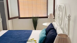A bedroom with a bed, a bedside table, a lamp, a window with blinds, and a mirror. - La Caracola (Benicarlo)