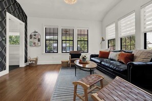 A spacious living room with a black leather sofa, a wooden bench, and a coffee table. There are three large windows with black frames and a chandelier hanging from the ceiling. - Creek's Edge Peaceful Retreat with Firepit (Fredericksburg)