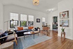 A spacious living room with a black leather sofa, a wooden coffee table, and a striped rug. There are wicker chairs and a dining area with a table and chairs.