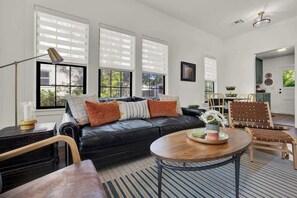 A living room with a black leather sofa, wooden chairs, a round coffee table, and large windows with blinds. - Creek's Edge Peaceful Retreat with Firepit (Fredericksburg)