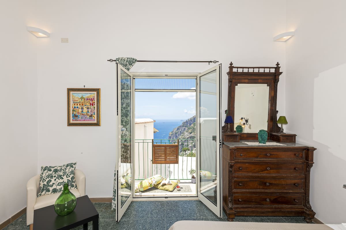 A room with a balcony, a wooden dresser, a framed picture, and a green vase on a table.
