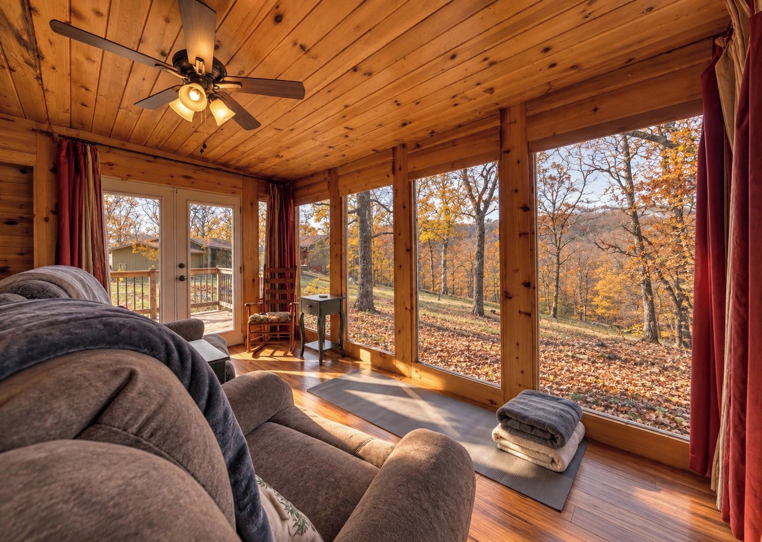 A cozy cabin interior with a wooden ceiling, a ceiling fan, a rocking chair, a small table, and a view of a forest through large windows.