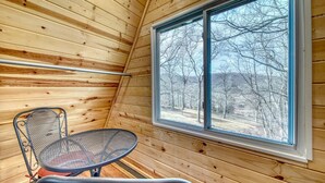 L’intérieur d’un chalet en bois, avec une table et une chaise en métal, et une grande fenêtre donnant sur les arbres.
