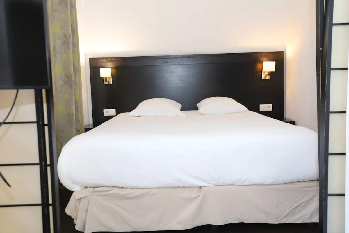 A neatly made bed with white linens, a dark headboard, and two wall-mounted reading lights.
