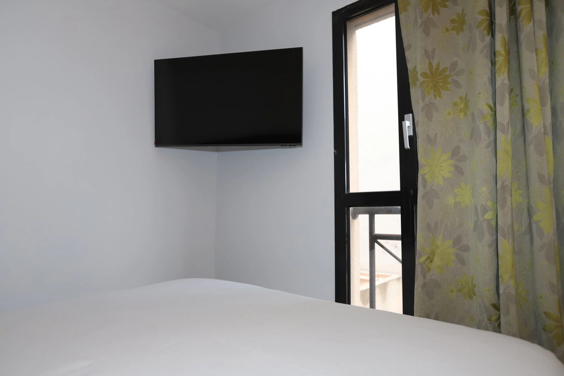 A room with a bed, a flat-screen TV mounted on the wall, and a window with floral curtains.