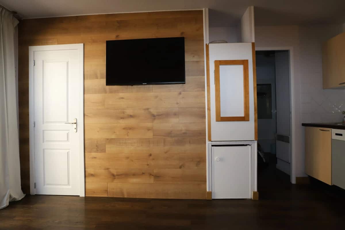 A room with a wooden wall, a white door, a flat-screen TV mounted above it, and a white refrigerator with a wooden frame.