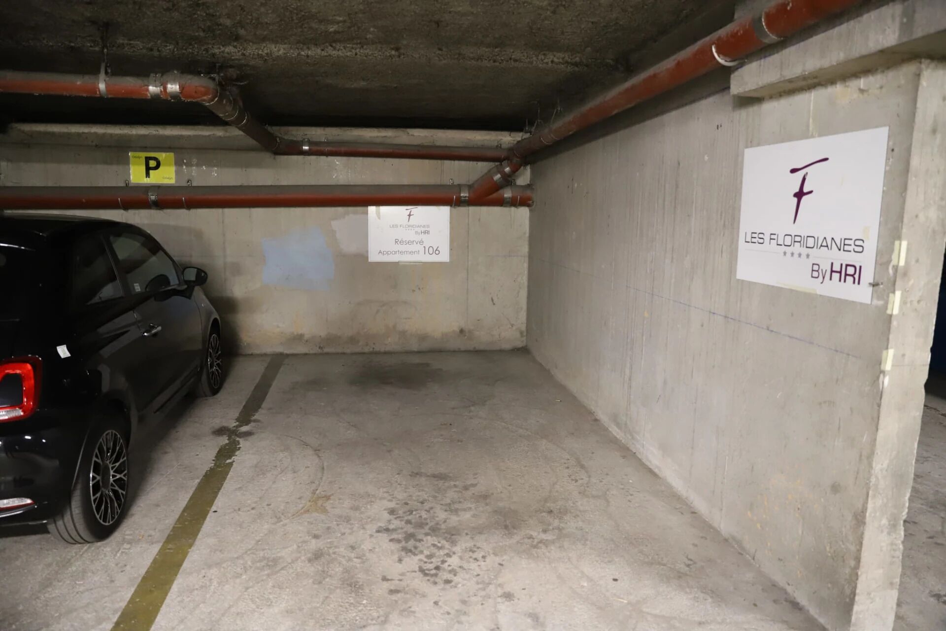Un parking avec une voiture noire garée sur une place signalée par un « P » jaune. Les murs sont en béton et le plafond laisse apparaître des tuyaux.
