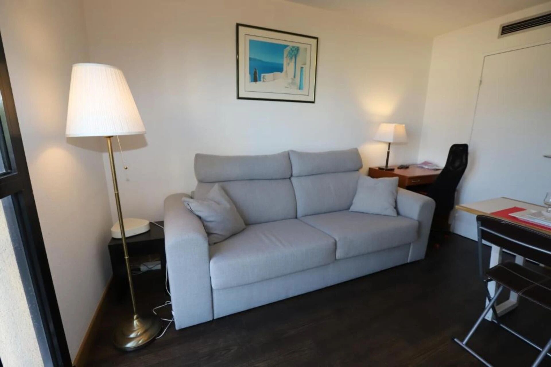A modern room with a grey sofa, two floor lamps, a desk with a chair, and a framed picture on the wall.