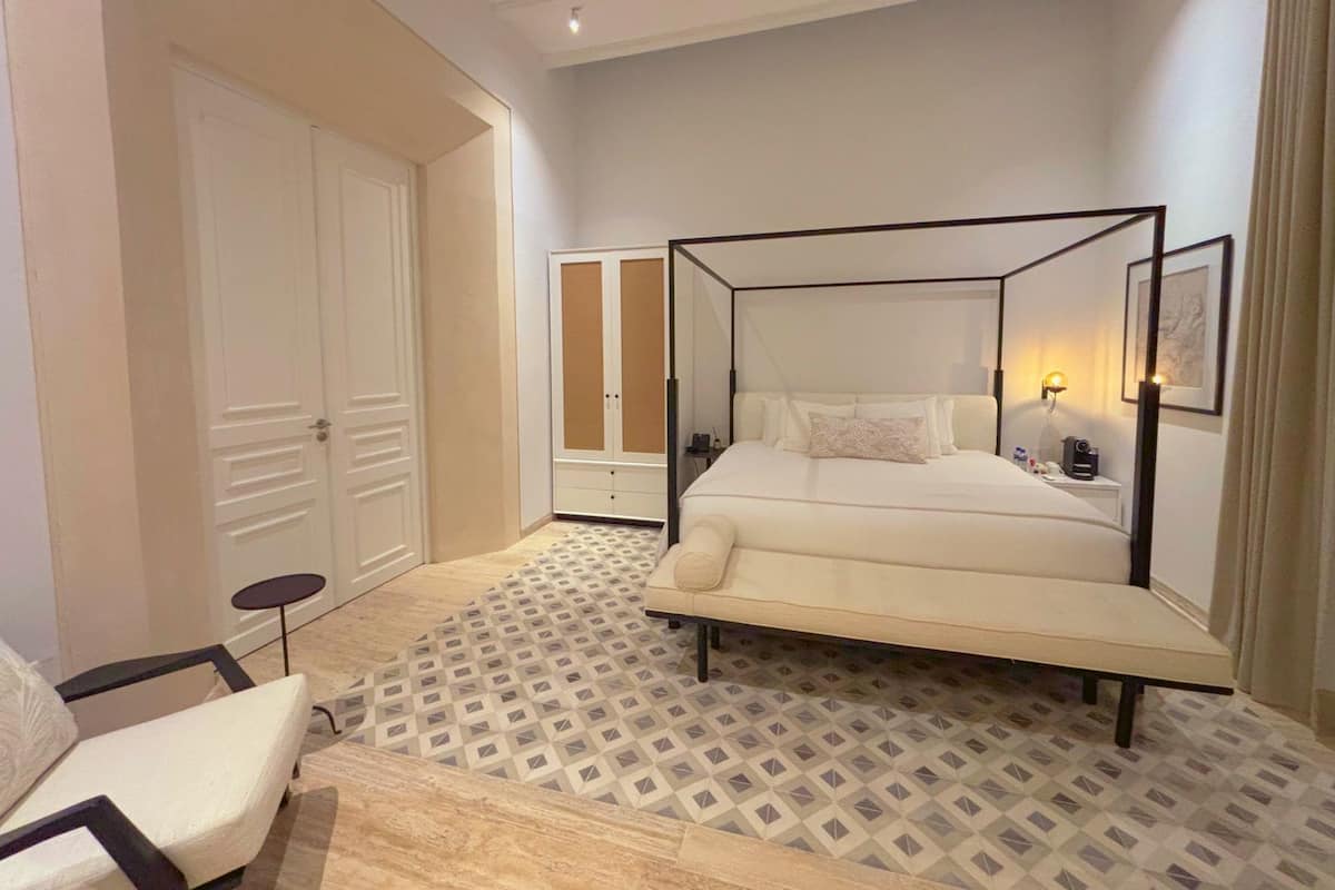 Een slaapkamer met een hemelbed, nachtkastje, stoel, kledingkast en een vloer met patroon.