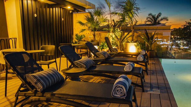 Un espace aménagé au bord de la piscine, avec des chaises longues et une table, entouré de plantes et d’une terrasse en bois.