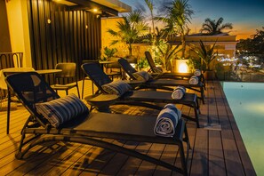 Un área junto a la piscina con sillones de descanso y una mesa, rodeada de plantas y una terraza de madera.