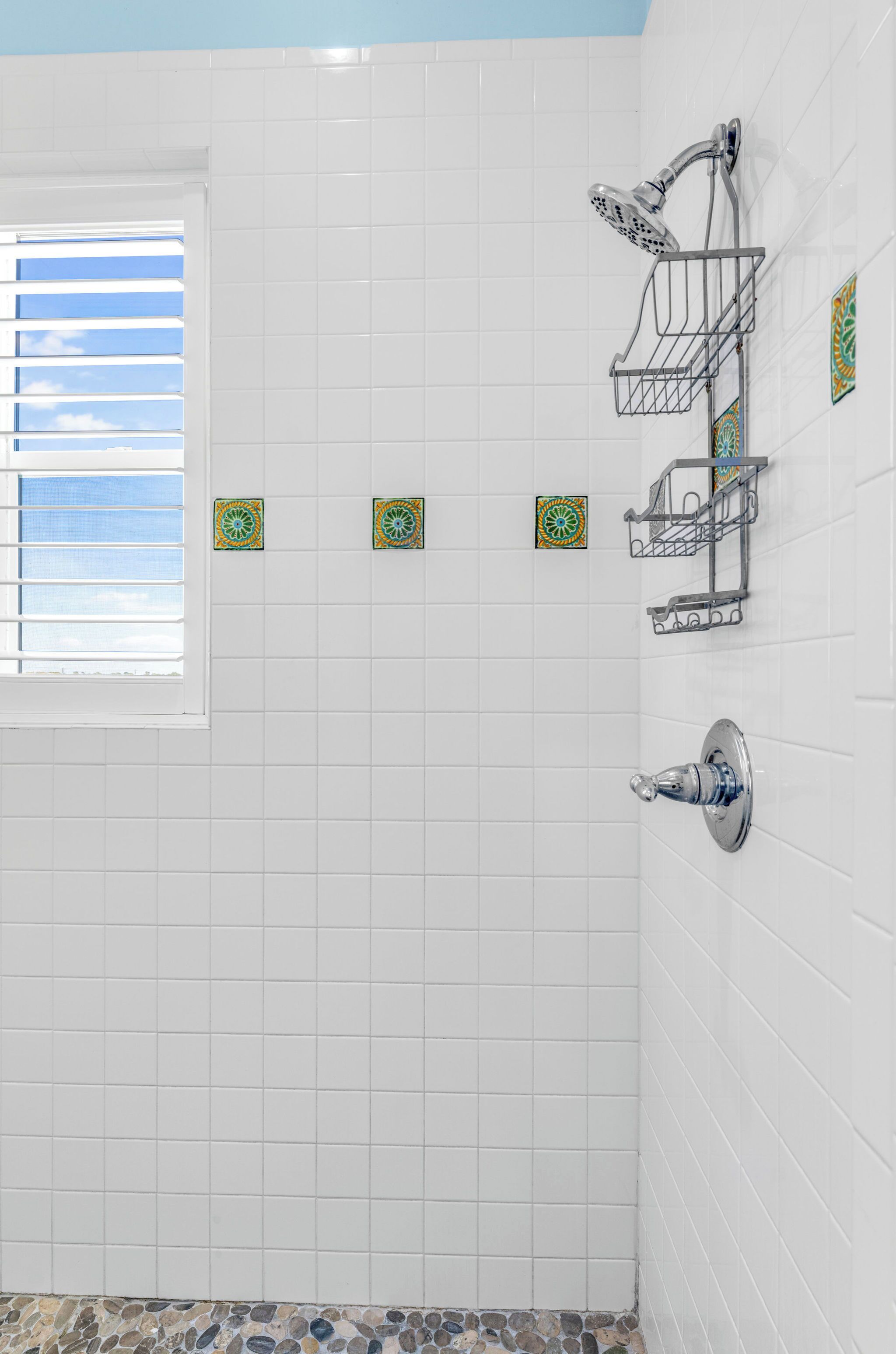 A bathroom with white tiled walls, a shower with a chrome faucet, a window with blinds, and decorative tiles on the wall.