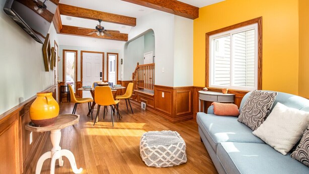 A spacious living area with a wooden ceiling, a dining table with chairs, a sofa with cushions, and a large window with blinds.