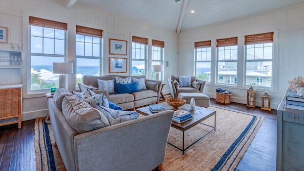 A spacious living room with a large sectional sofa, a coffee table, and a ceiling fan. There are multiple windows with blinds, and a view of the ocean is visible through the windows. - Sky High • Panoramic Views, Dock Slip, Steps to Tahiti Beach - Hope Town (Hope Town)