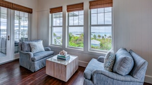 A spacious room with a large window offering a view of the ocean and a balcony. There are two upholstered chairs and a small coffee table with books. - Sky High • Panoramic Views, Dock Slip, Steps to Tahiti Beach - Hope Town (Hope Town)