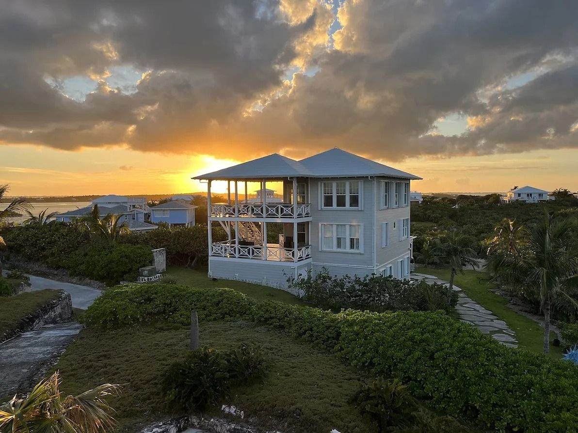 A two-story house with a balcony, surrounded by lush greenery and a view of the sunset.