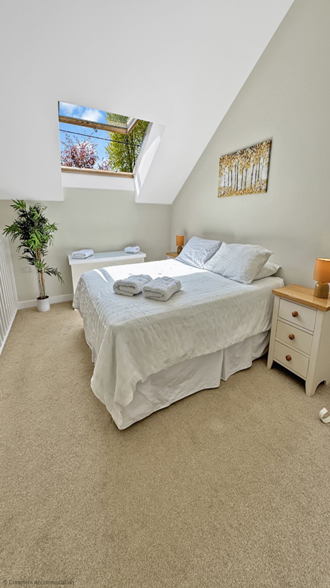 Quarto com claraboia, cama com roupa de cama branca, mesa de cabeceira com abajur e uma planta em vaso.