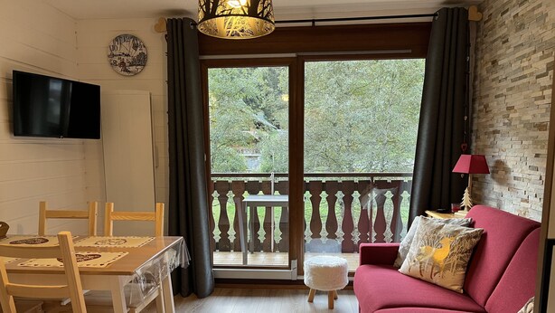 A living room with a red sofa, a wooden dining table with chairs, a TV mounted on the wall, and a balcony with a view of trees. - Appartement Pied des Pistes (Châtel)