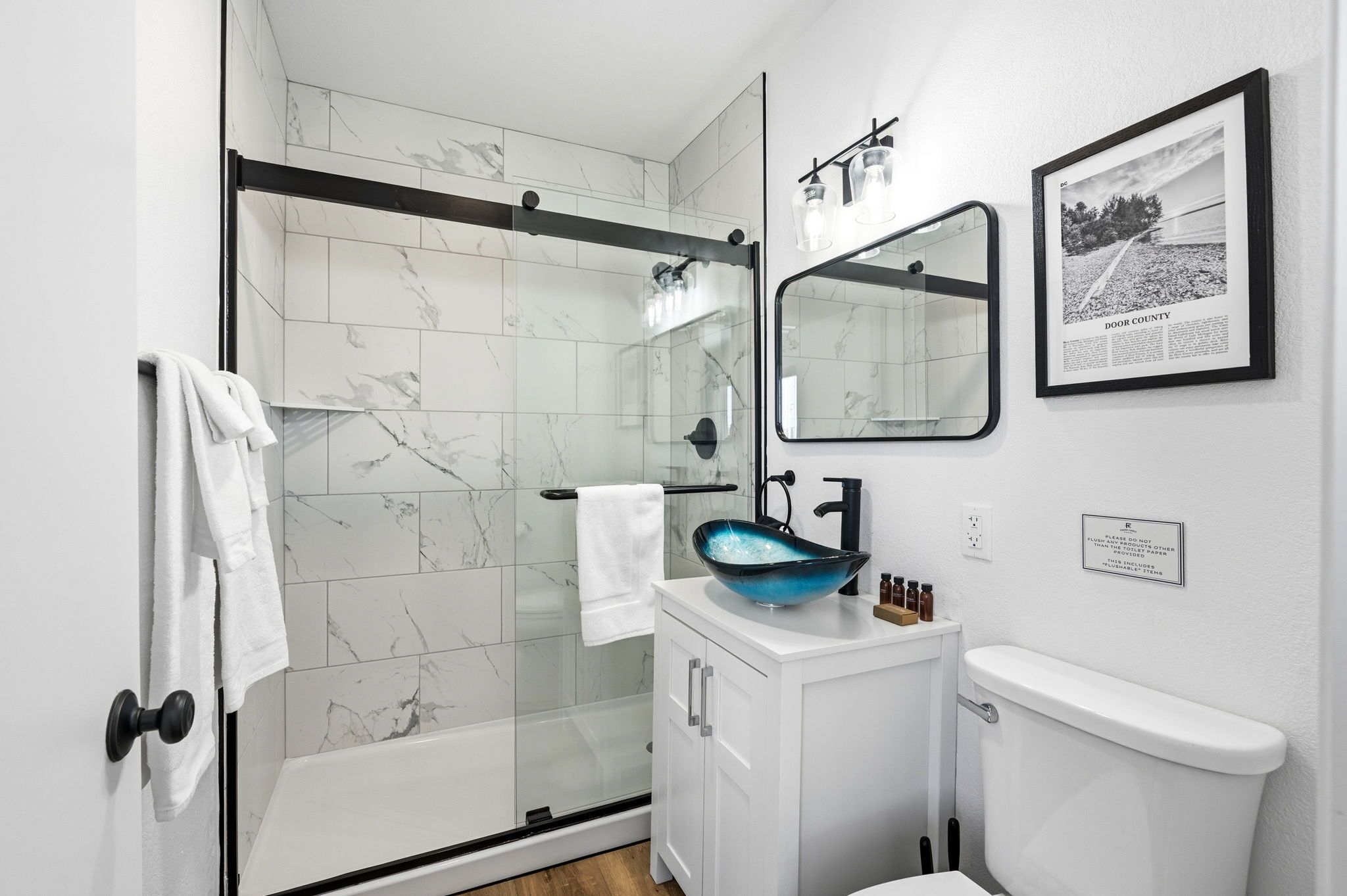 Une salle de bain moderne avec une douche à parois de verre, un meuble-lavabo blanc doté d’un vasque bleue et une photographie encadrée accrochée au mur.