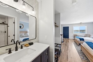 Uma casa de banho moderna com um espelho grande, uma sanita e um chuveiro. O quarto tem soalho em madeira e vista para a praia através de uma janela.