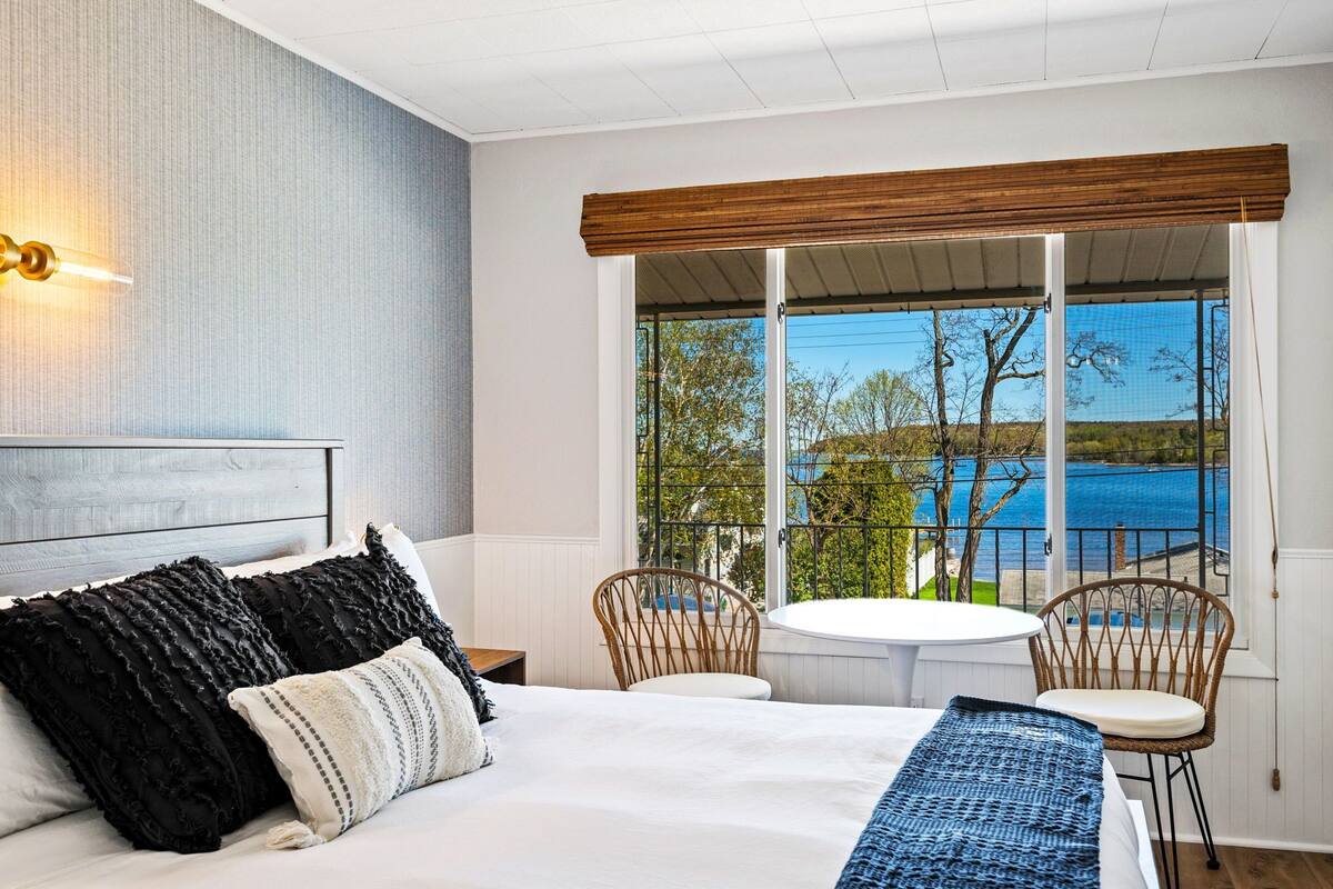 A bedroom with a large bed, two chairs, a small table, and a view of a lake.