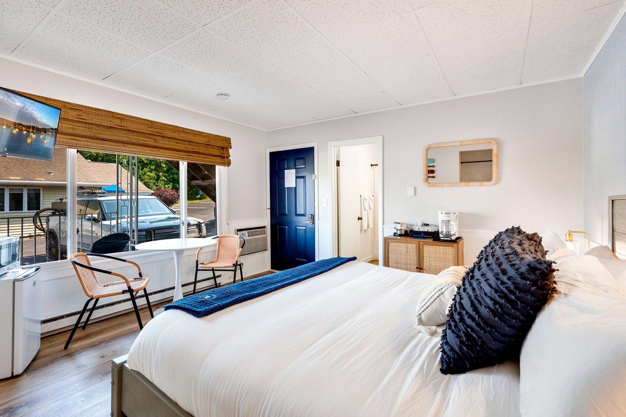 Een hotelkamer met een bed, een bureau, een stoel, een raam met uitzicht op een auto en bomen, en een deur.