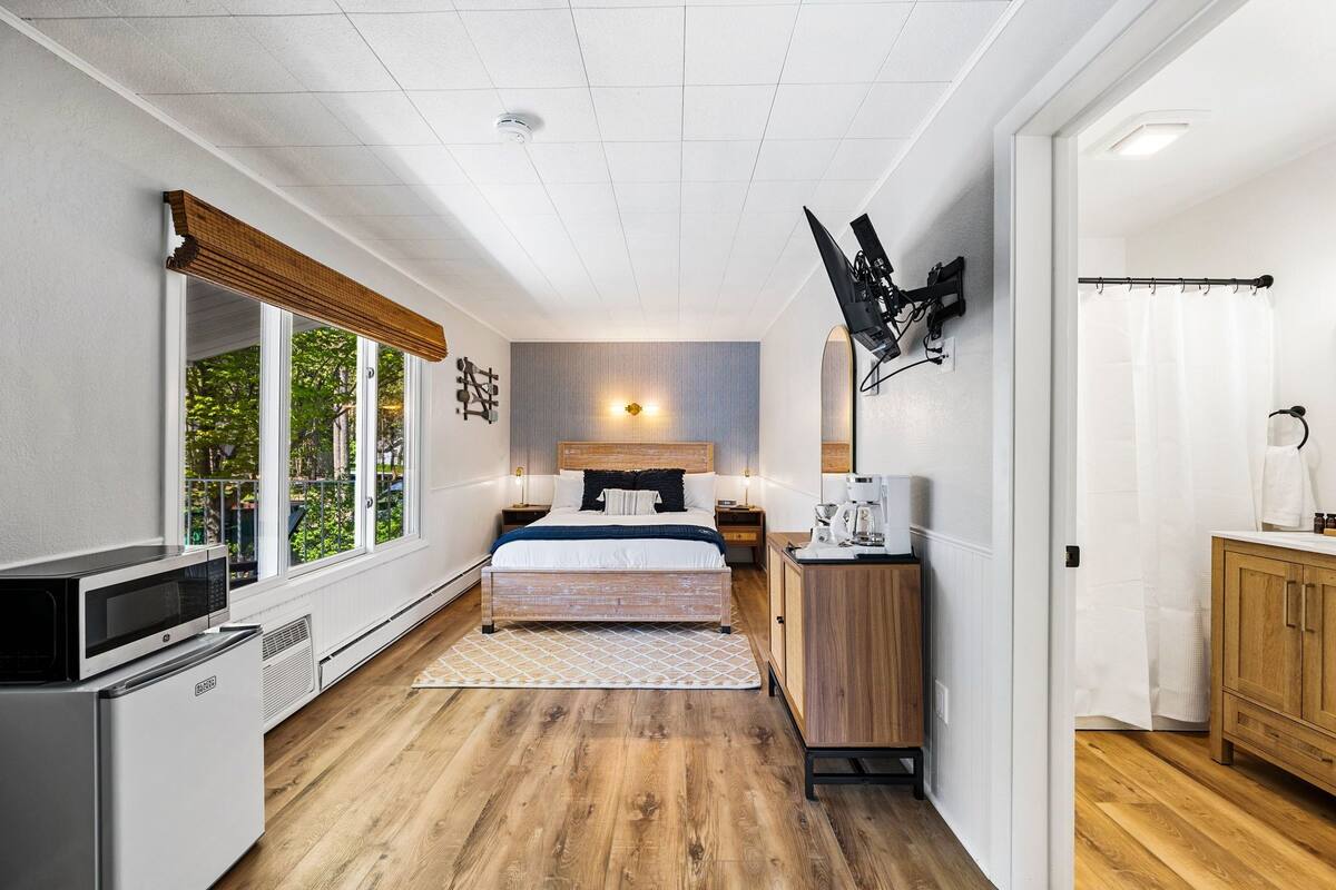 A modern bedroom with a bed, bedside tables, a microwave, and a view of greenery through large windows.
