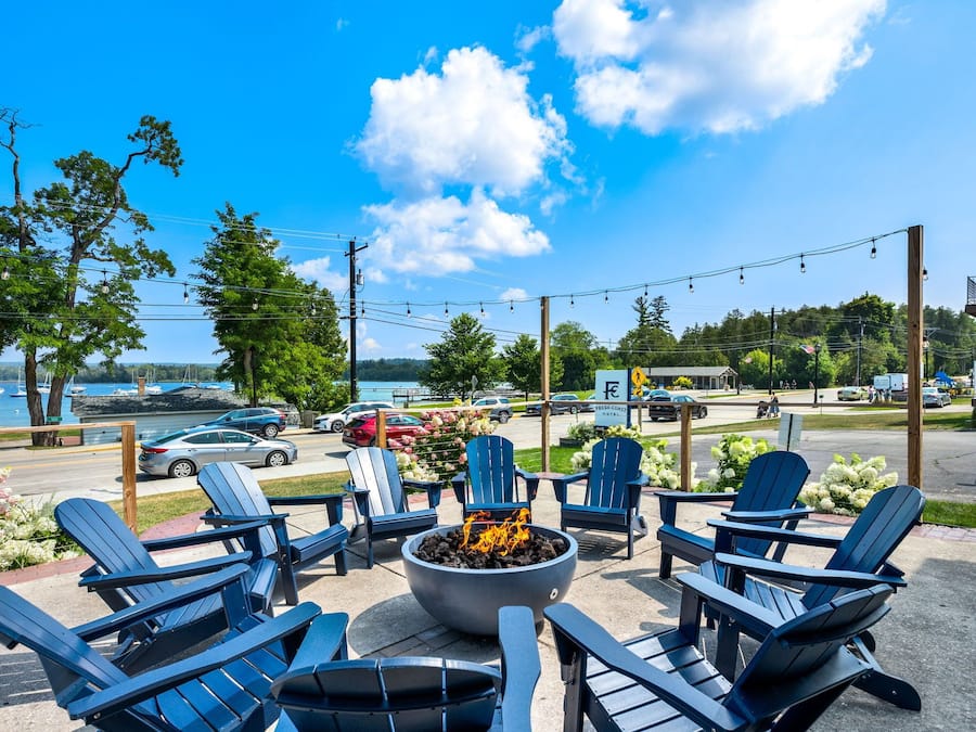 An outdoor seating area with Adirondack chairs and a fire pit, overlooking a body of water and a road with vehicles.