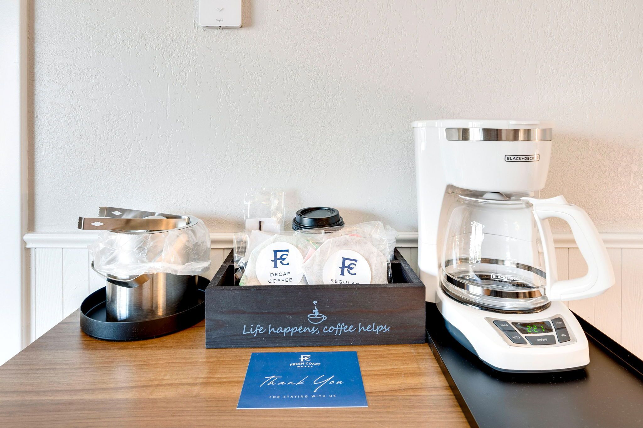 A coffee station with a coffee maker, a glass carafe, and a box of coffee pods on a wooden table.