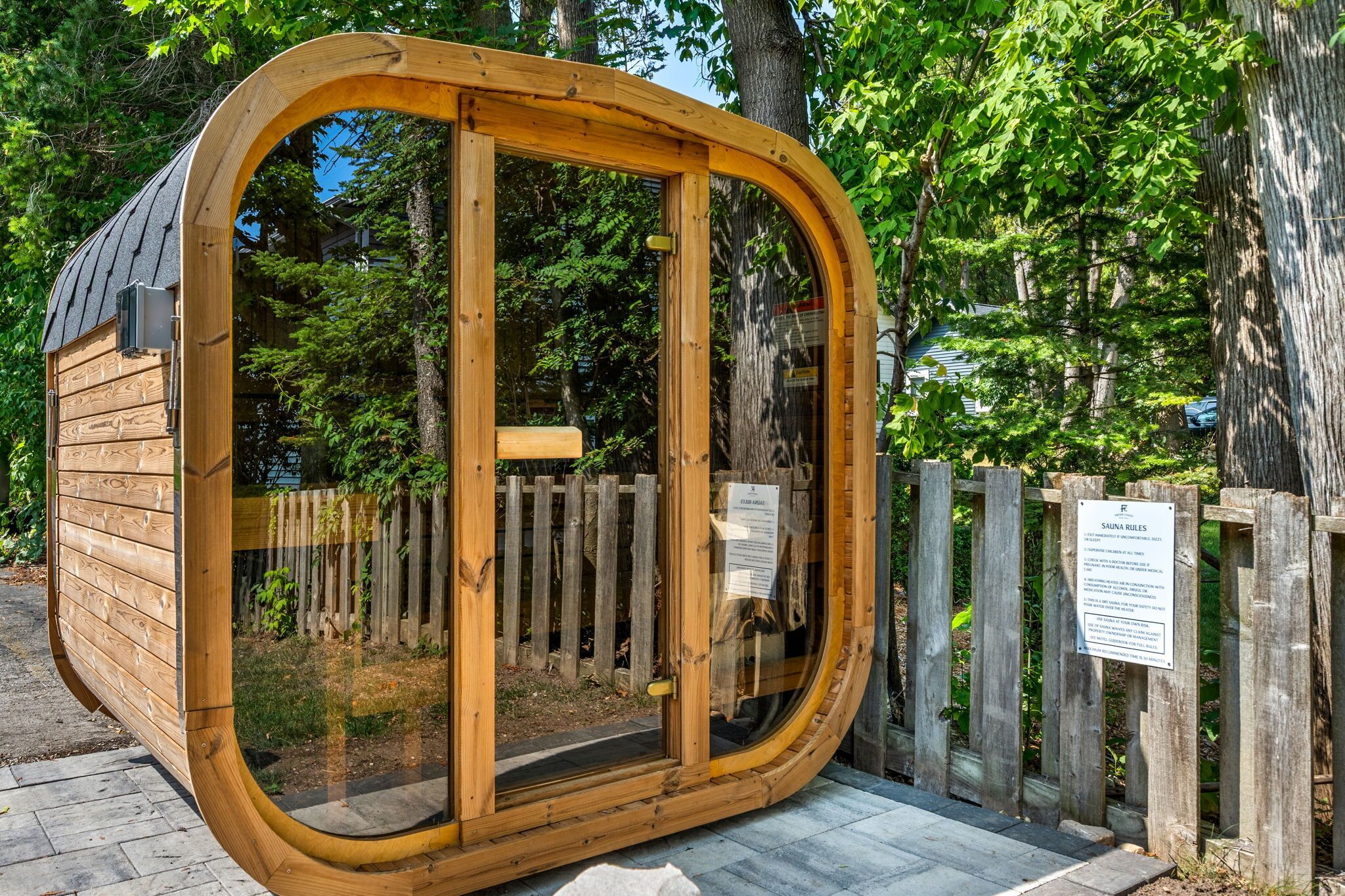 A wooden sauna structure with a glass door, surrounded by a wooden fence and trees.