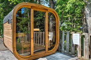 A wooden sauna structure with a glass door, surrounded by a wooden fence and trees. - Fresh Coast Motel (Fish Creek)