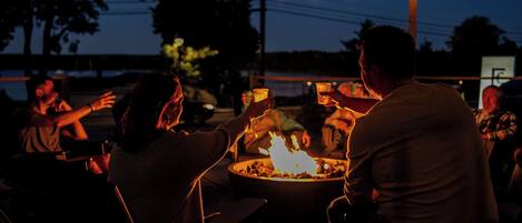 A group of people seated around a table with a fire, under string lights.