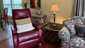 A living room with a red leather armchair, a patterned sofa, a glass coffee table, a lamp, a painting, and a vase with flowers. - BestStays@Bayou-Chic -work crews love us. Fenced with WIFI (Baton Rouge)