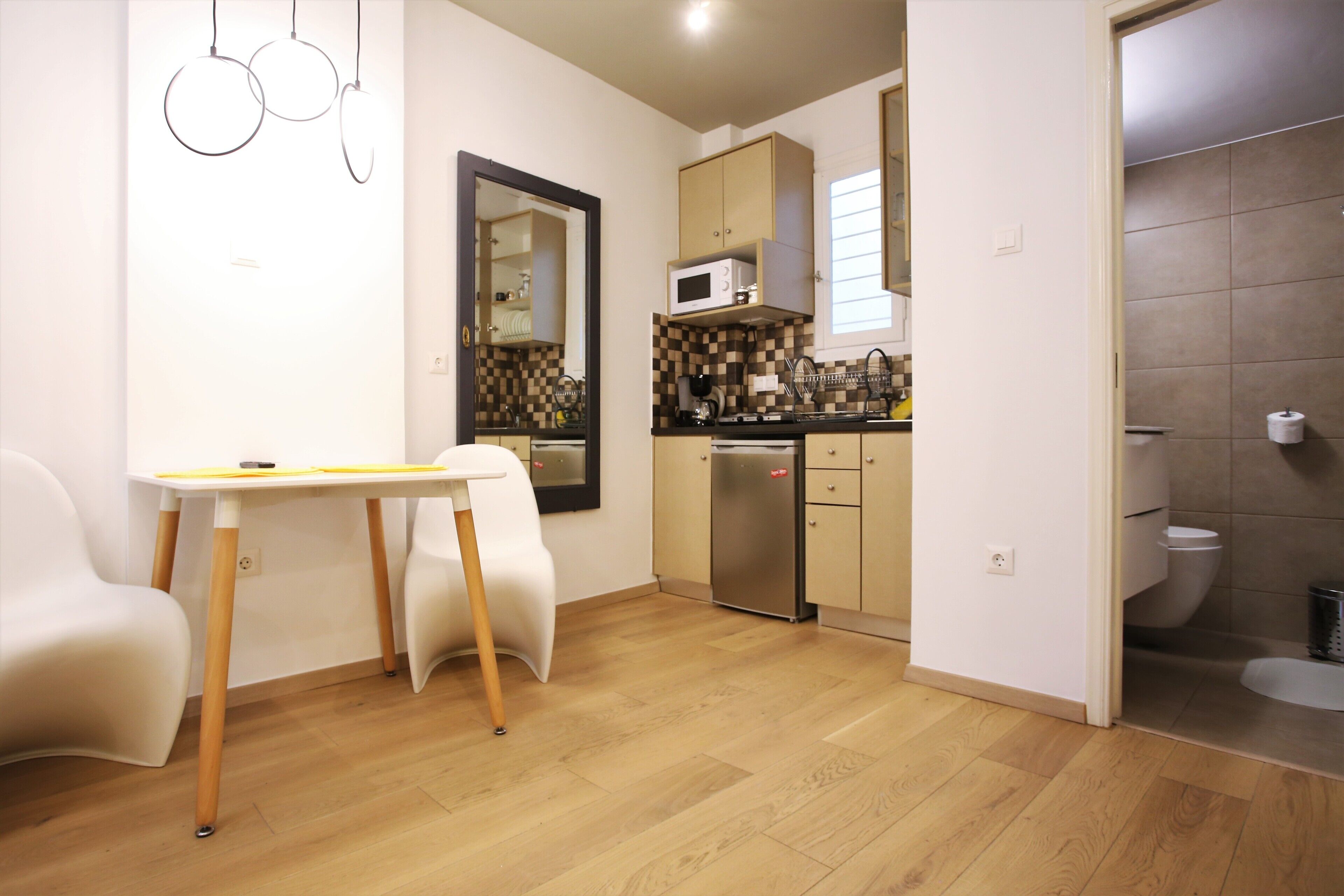 A compact kitchen with a microwave, sink, and dishwasher. There is a dining table with chairs and a mirror on the wall. The room has wooden flooring and a tiled shower area.