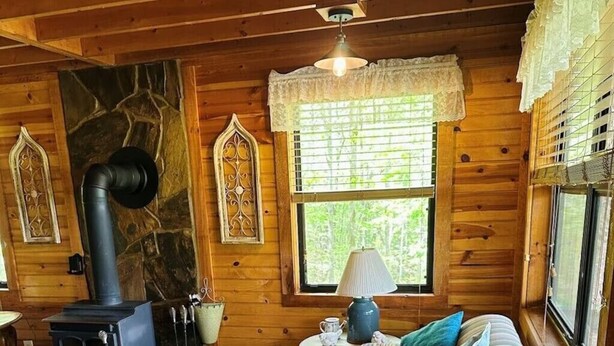 Ein gemütliches Blockhaus-Interieur mit Holzvertäfelung, einem Kamin aus Naturstein, einem gestreiften Sofa und einem Fenster mit Spitzenvorhängen.