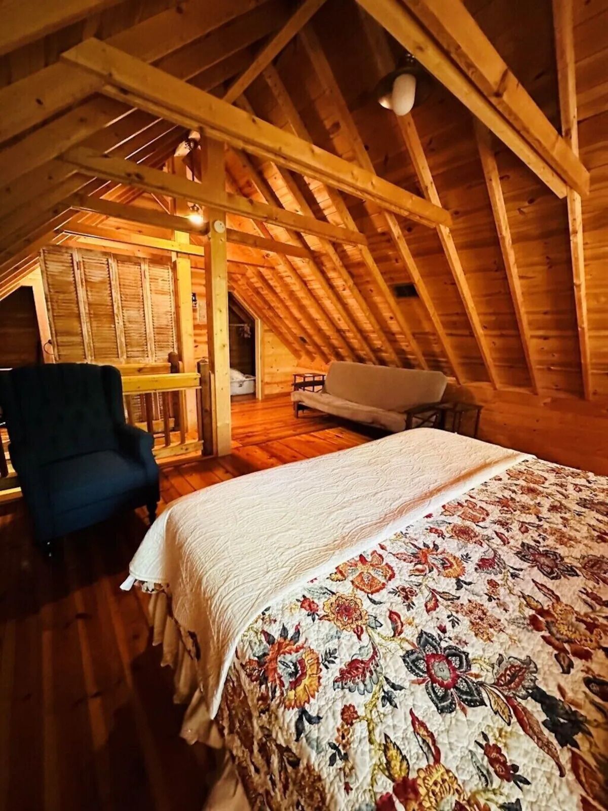 A cozy attic bedroom with a wooden ceiling, a bed with a floral bedspread, a blue armchair, and a small sofa.