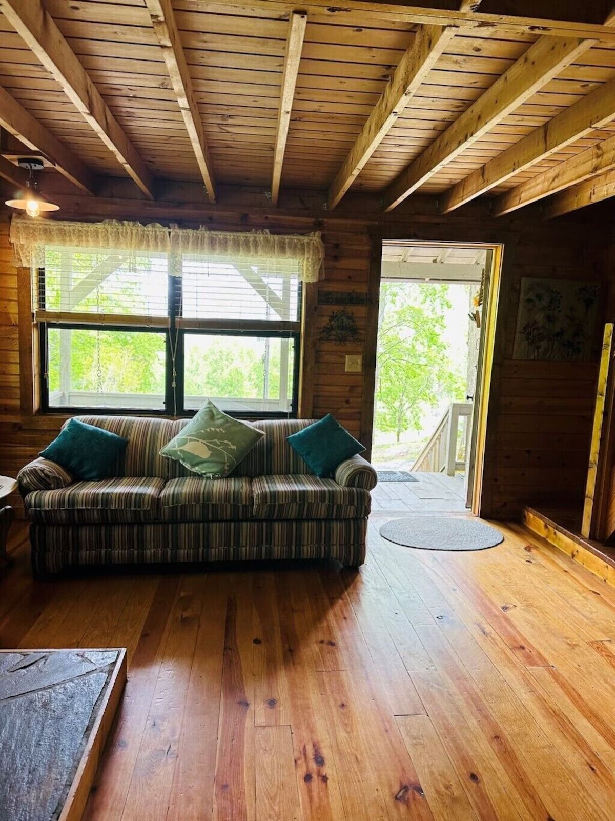 A wooden cabin interior with a striped sofa, a wooden floor, and a window with a view of greenery.