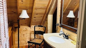 Ein kleines Badezimmer aus Holz mit schrägverlaufender Decke, einem einzelnen Stuhl, einem Waschbecken und zwei Stehlampen.