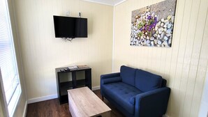 Ein Wohnzimmer mit einem blauen Sofa, einem Holztisch, einer schwarzen TV-Konsole und einem Bild von Steinen und Blumen an der Wand.