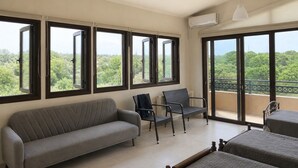 A room with a sofa, two chairs, a bed, large windows, and an air conditioning unit.