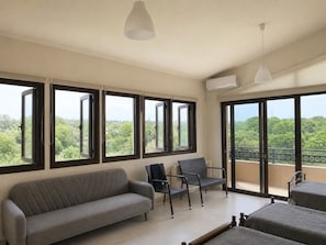 A room with a sofa, two chairs, a bed, large windows, and an air conditioning unit.